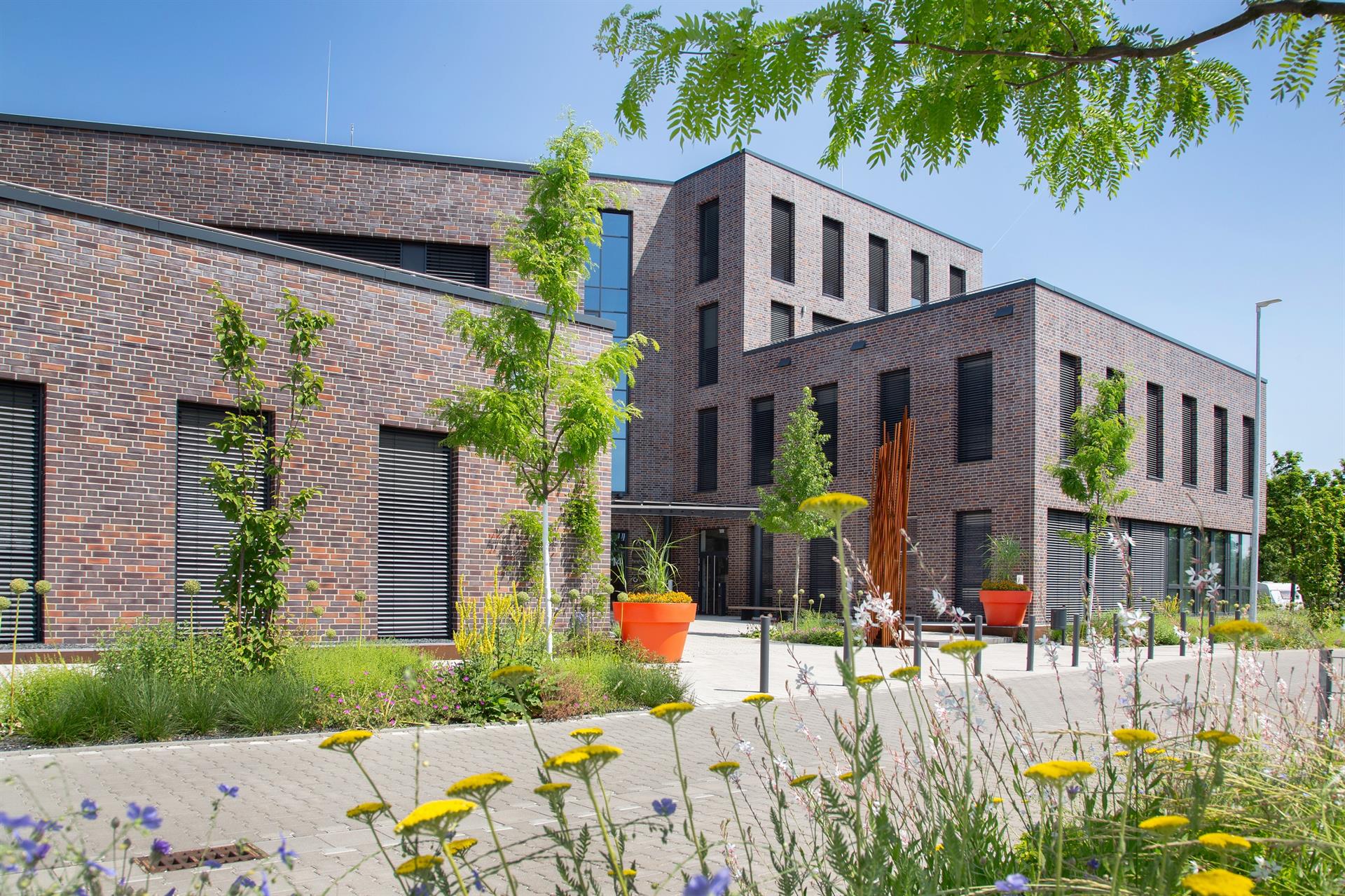 Bürogebäude bei blauem Himmel mit grünen Bäumen und bunten Blumen im Vordergrund.